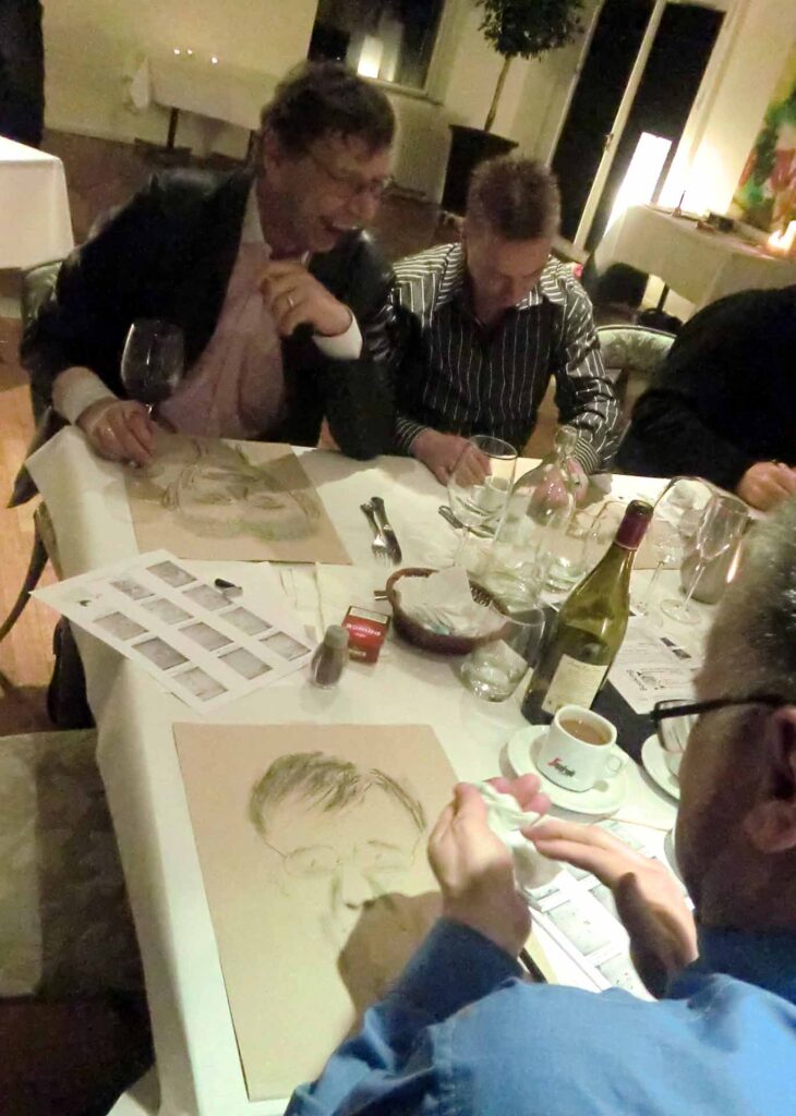 Tre grinende og tegnende m?nd er fotograferet under en Karikaturworskshop ved et uformelt bord med vin, glas, cigaretter og papir. Man ser svagt den ene mands karikaturtegning.